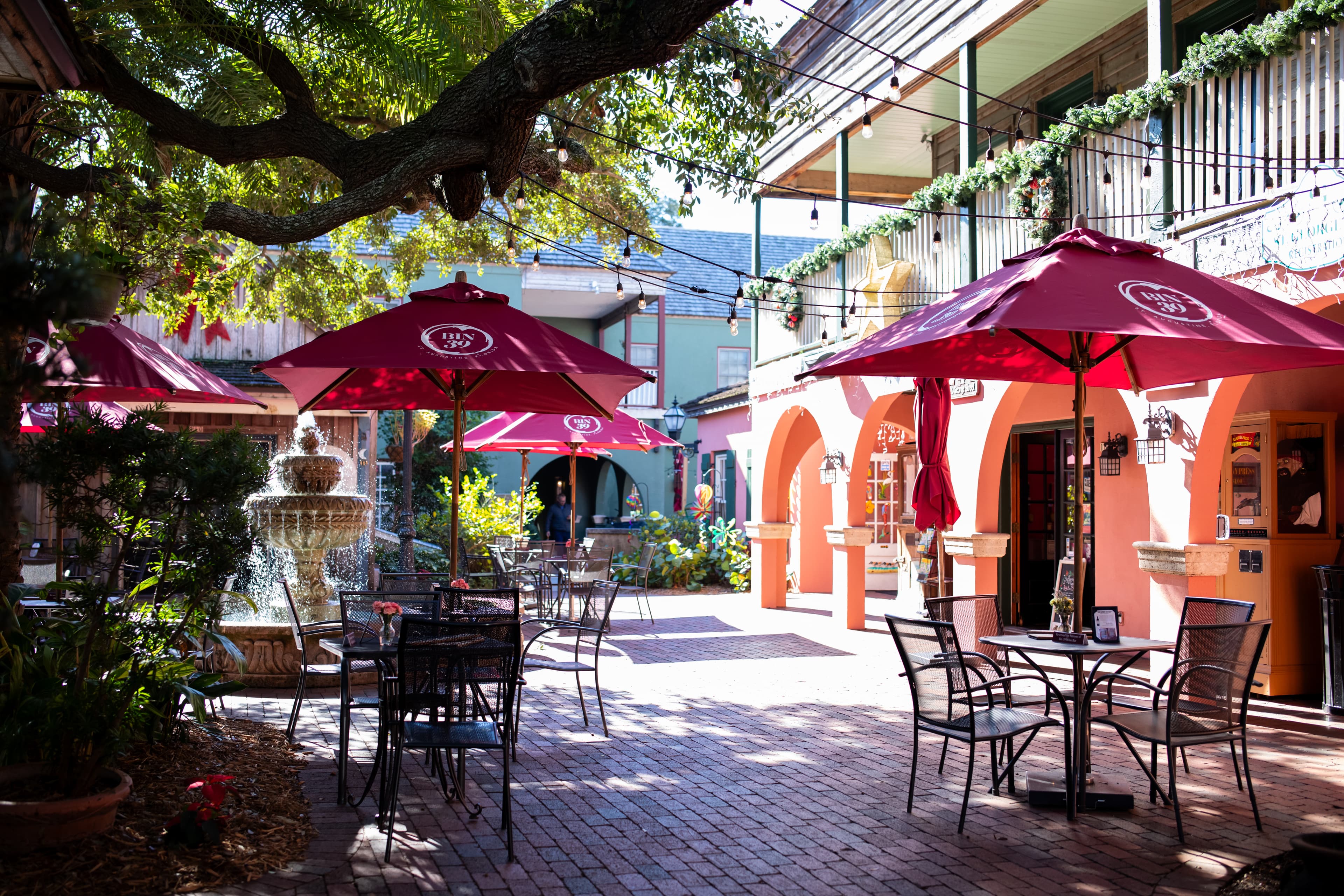 A charming outdoor dining area with red umbrellas, tables, and a fountain amidst lush greenery.