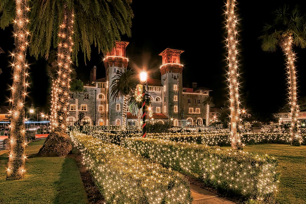 A historic building adorned with festive lights surrounded by landscaped greenery at night.