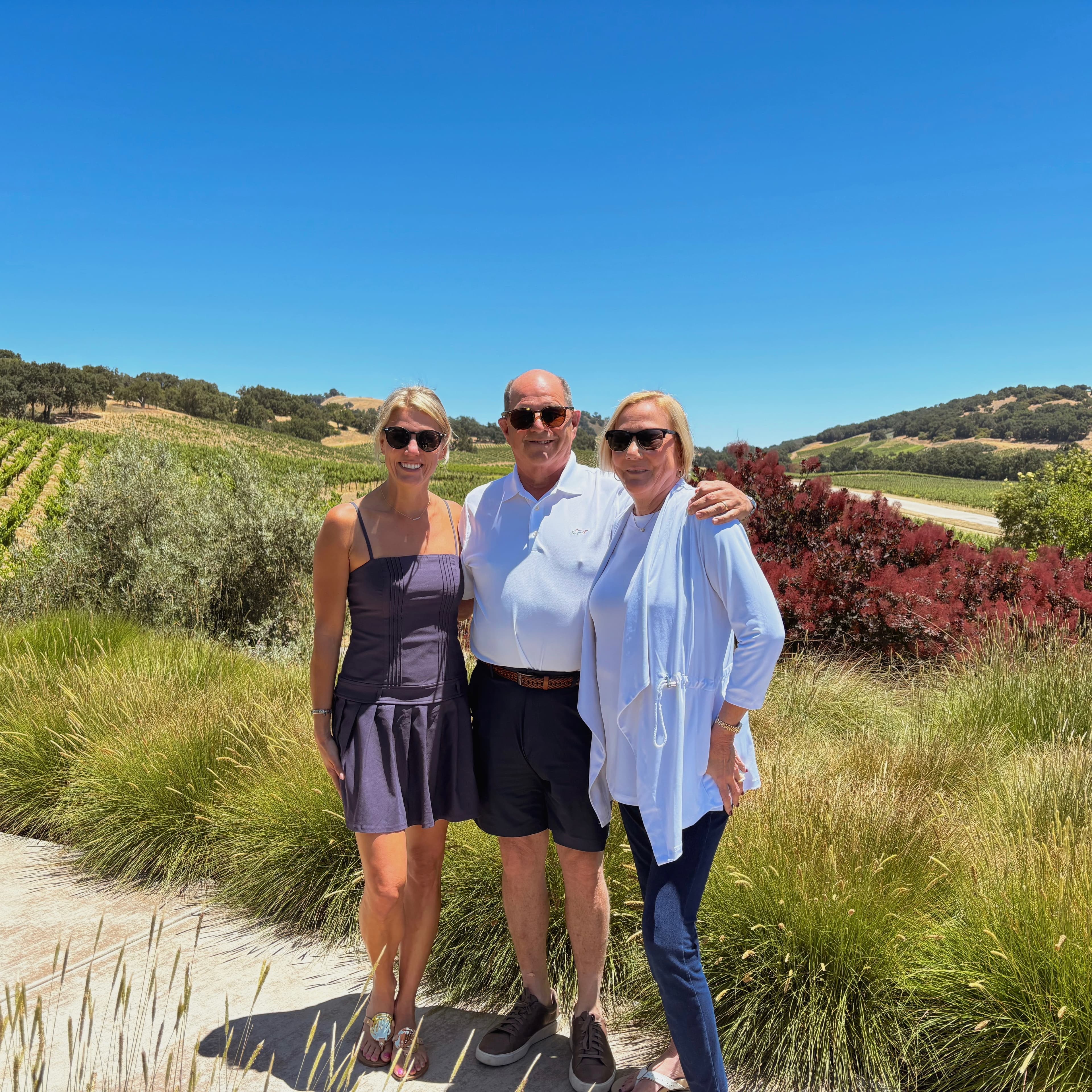 Three people pose together in a sunny vineyard setting, surrounded by lush greenery.