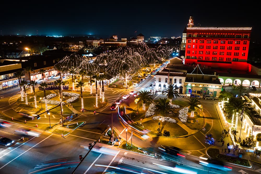 Aerial view of a circular plaza illuminated with holiday lights at night, featuring palm trees and a historic building.
