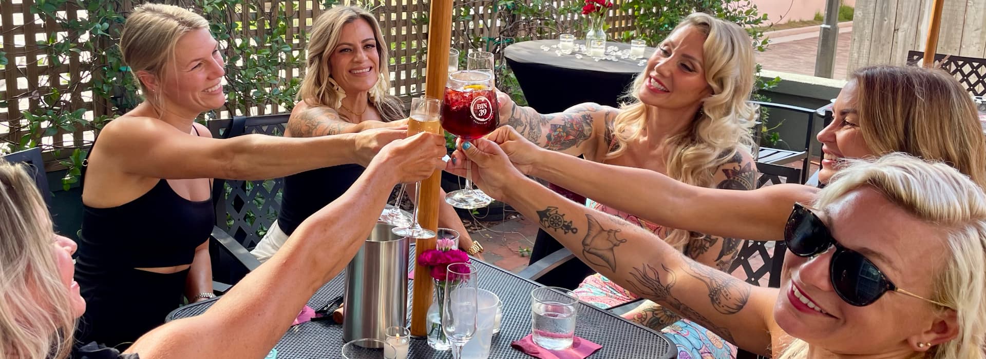 A group of six women clinks glasses together in celebration at an outdoor table.