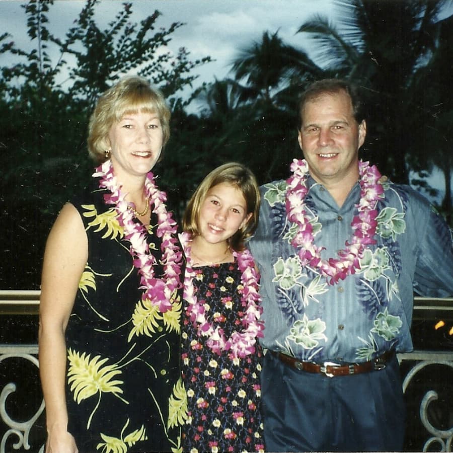 A smiling woman, a girl, and a man wear flower leis against a tropical backdrop.