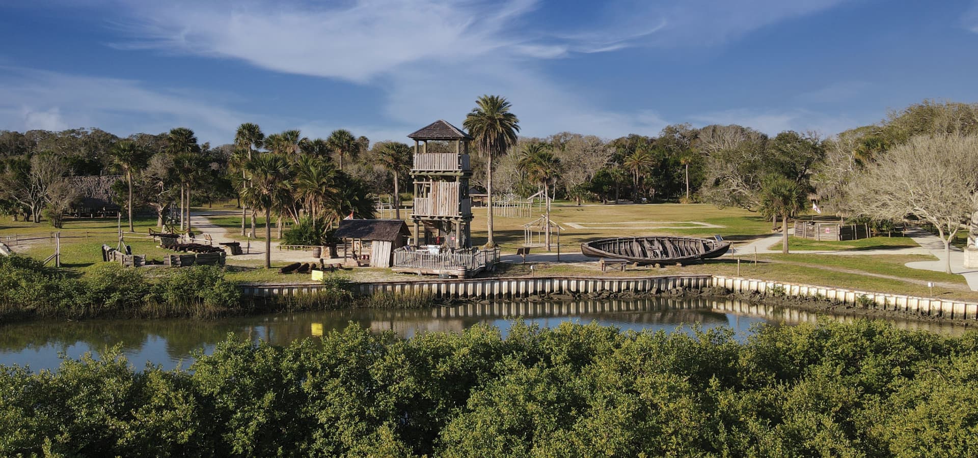 A scenic view of a playground with a tall wooden structure, surrounded by palm trees and a calm waterway.