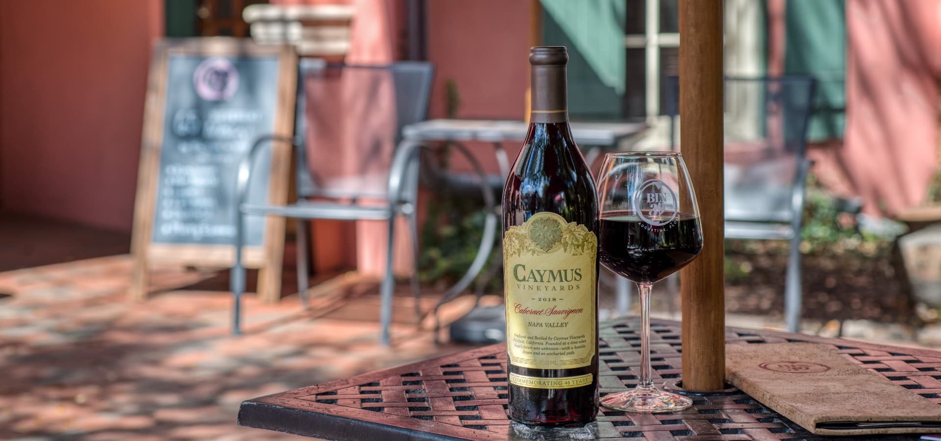 A bottle of Caymus Vineyards Cabernet Sauvignon and a glass of red wine sit on a table in a cozy outdoor setting.