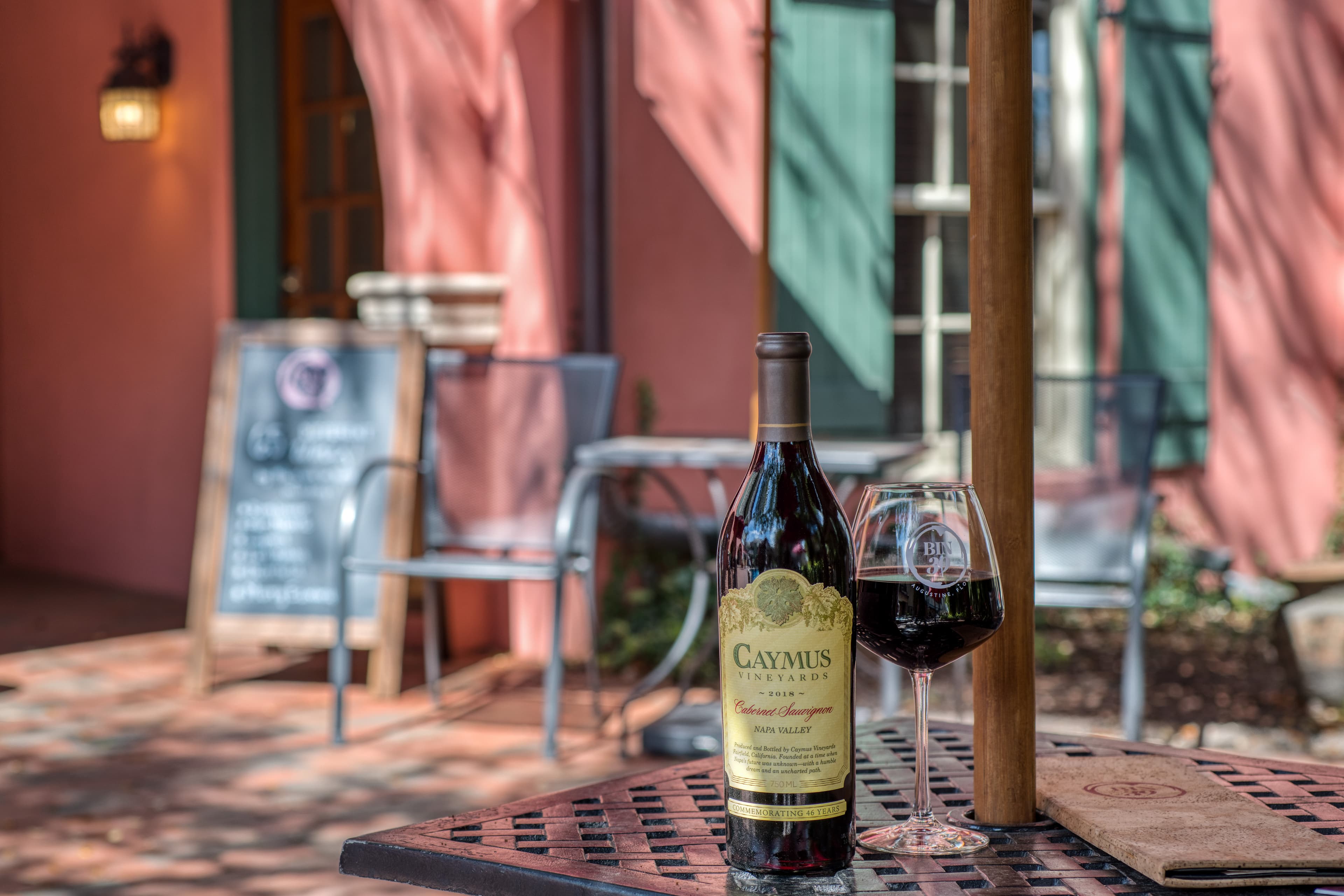 A bottle of Caymus Cabernet Sauvignon and a glass of red wine sit on a table at an outdoor café.