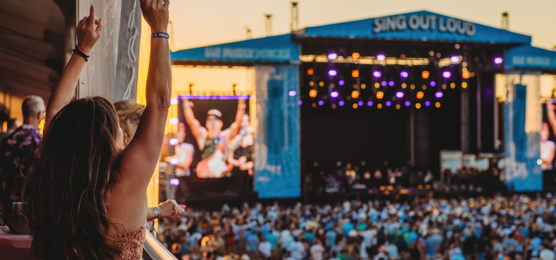 A concertgoer with arms raised enjoys a live performance at sunset.