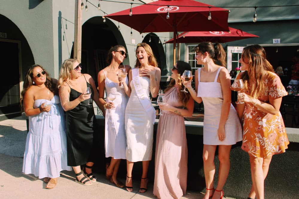 A group of seven women celebrating together outdoors, holding drinks and laughing.