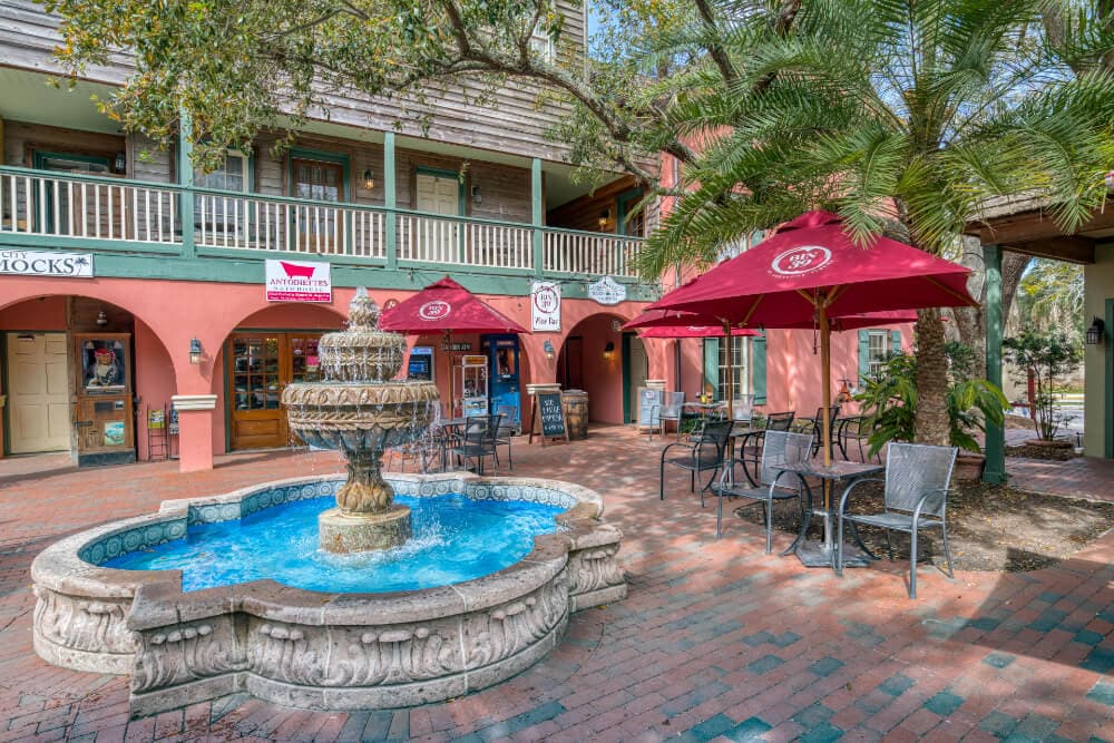 A decorative fountain surrounded by outdoor seating and shops under patio umbrellas.
