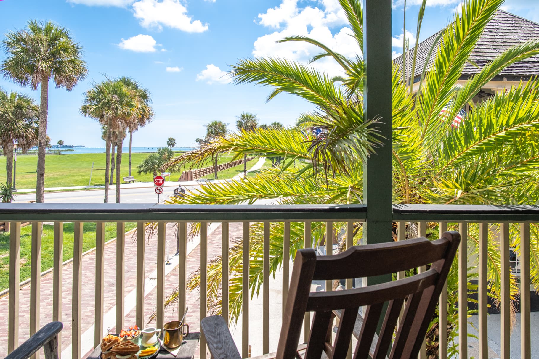 View from a balcony featuring palm trees, a rocking chair, and a glimpse of the ocean under a blue sky.